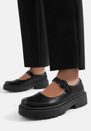 Černé kožené boty Mary Jane s robustní pryžovou podešví a kulatou špičkou. Mají přezkový pásek a plochý podpatek. Texturovaný vzor podrážky.