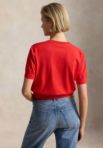 Rövid szőke hajú nő piros rövid ujjú pulóvert és kék farmernadrágot visel, háttal áll a kamerának.