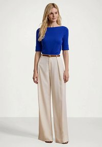 Sieviete stāv, ģērbusies zila elkoņu piedurkņu blūzē, ieslietā bēšās platās biksēs ar brūnu jostu un atvērtu purngalu apaviem.