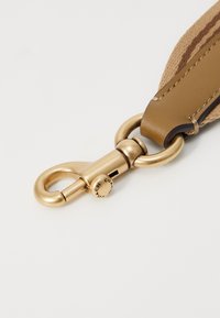 Mousqueton pivotant en métal doré fixé à une lanière en cuir fauve avec des détails en tissu tissé beige.