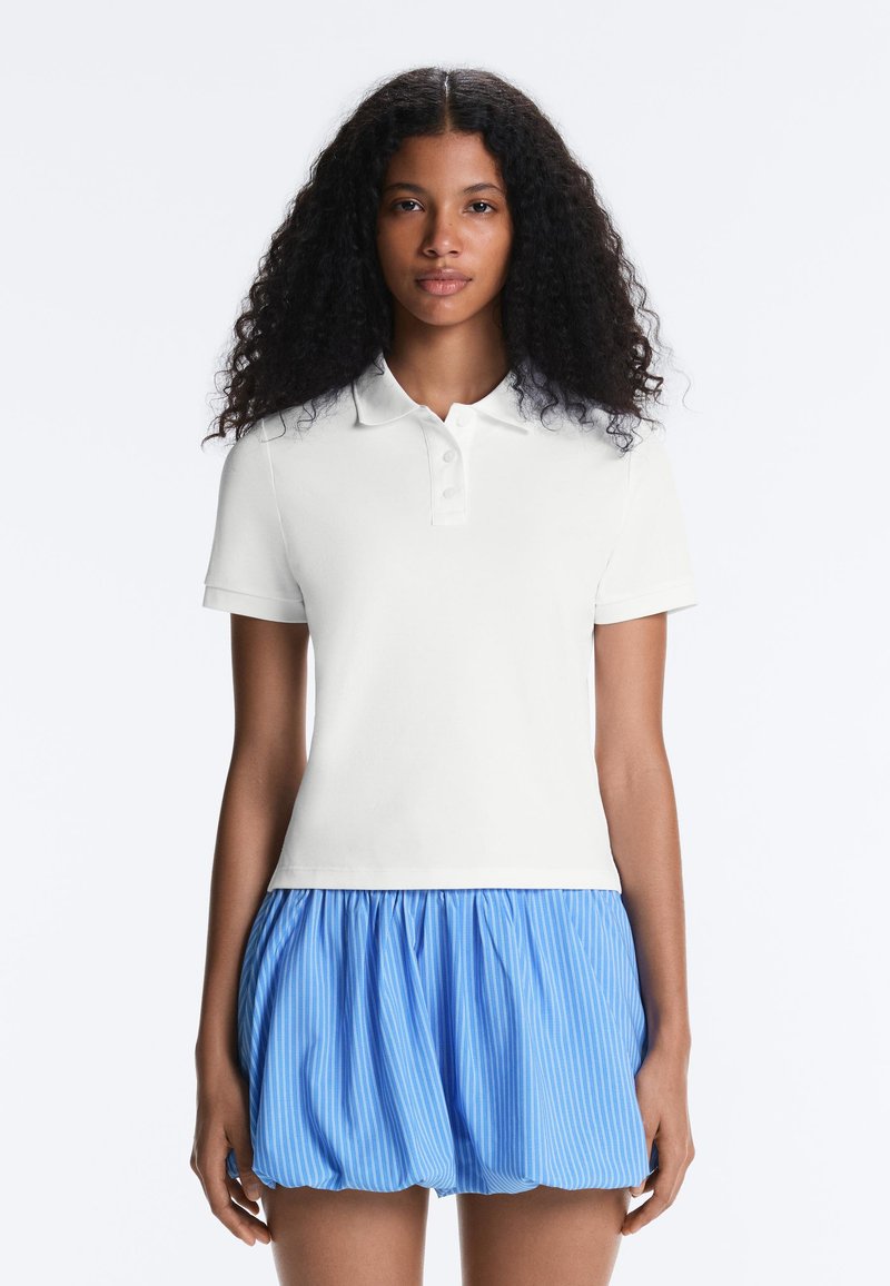 Femme aux cheveux bouclés portant un polo blanc à manches courtes et une mini-jupe bleue rayée froncée, debout devant un fond blanc.