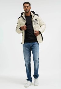 Man wearing a beige and black hooded jacket with text, black sweatshirt, blue jeans, and white sneakers, adjusting jacket zipper.