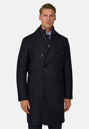 Homme portant un pardessus noir croisé avec de larges revers par-dessus une couche intérieure zippée et une cravate, debout devant un fond uni.