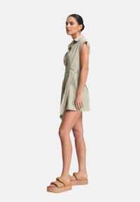 Abito corto verde chiaro con colletto a bottoni, vita cinturata e design senza maniche. Indossato con sandali platform beige con fasce larghe.