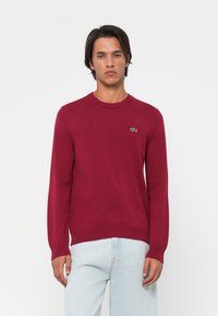Suéter de malha burgundy com decote redondo e mangas compridas. Apresenta um pequeno logótipo de crocodilo verde no lado esquerdo do peito. Textura suave.