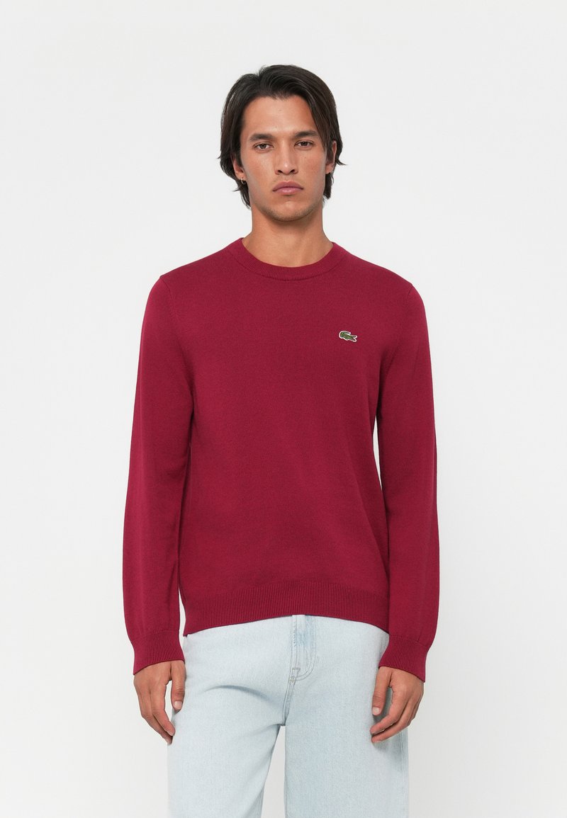 Suéter de malha burgundy com decote redondo e mangas compridas. Apresenta um pequeno logótipo de crocodilo verde no lado esquerdo do peito. Textura suave.