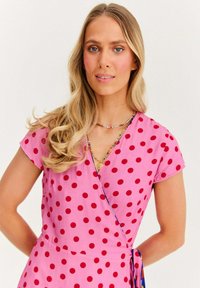 Růžové zavinovací šaty s výraznými červenými puntíky, krátkým rukávem a odpovídajícím pásem. Mají výstřih do V a lehkou, hladkou texturu látky.