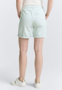 Shorts en coton vert clair avec une finition texturée, dotés de poignets roulés et de deux poches arrière. Associés à des baskets blanches avec des accents dorés.
