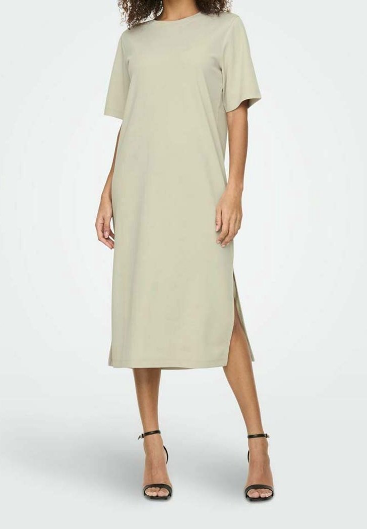 Femme portant une robe midi beige à manches courtes avec une fente latérale, associée à des sandales noires à talons hauts et bride à la cheville, debout sur un fond blanc.