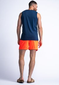 Camiseta sin mangas de color marino y bañador corto naranja brillante, fabricados con un tejido ligero, con un corte relajado y cinturilla plana. Sandalias marrones.