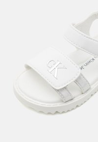 Biała sandałka z regulowanym paskiem na rzep, posiadająca okrągły obszar na palce i teksturowaną wkładkę. Zawiera srebrne logo.