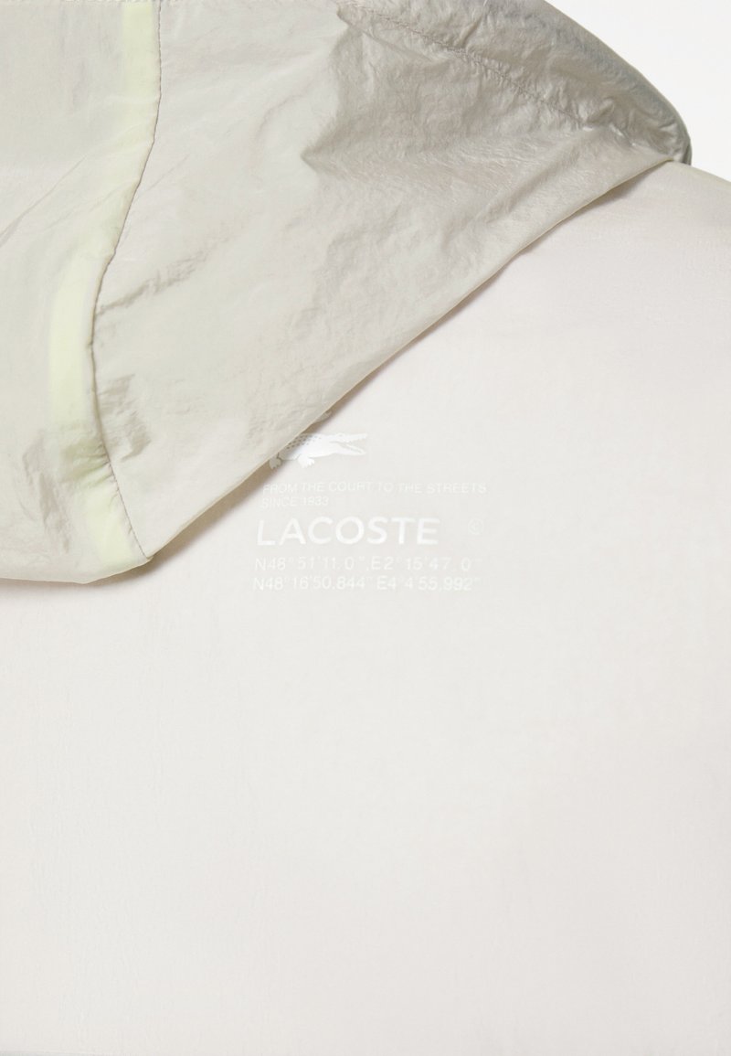 Lichtgrijze, met een capuchon, jas met een zachte, gladde textuur. Bevat een logo en geografische coördinaten op de borst; naadloos ontwerp.