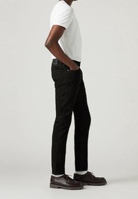 Pantalones de mezclilla negros, diseño ajustado, con bolsillos y una etiqueta de cuero en la cintura. Combinados con una camiseta blanca y zapatos marrones.