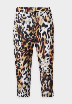 Mujer con camisa blanca de manga larga y cuello, pantalones cortos con estampado de leopardo y sandalias negras de tacón, de pie frente a un fondo liso.
