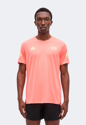 Άνδρας που φοράει κοραλλί T-shirt BMW Berlin Marathon με κίτρινα λογότυπα και μαύρα σορτς Adidas, στέκεται μπροστά σε απλό φωτεινό φόντο.