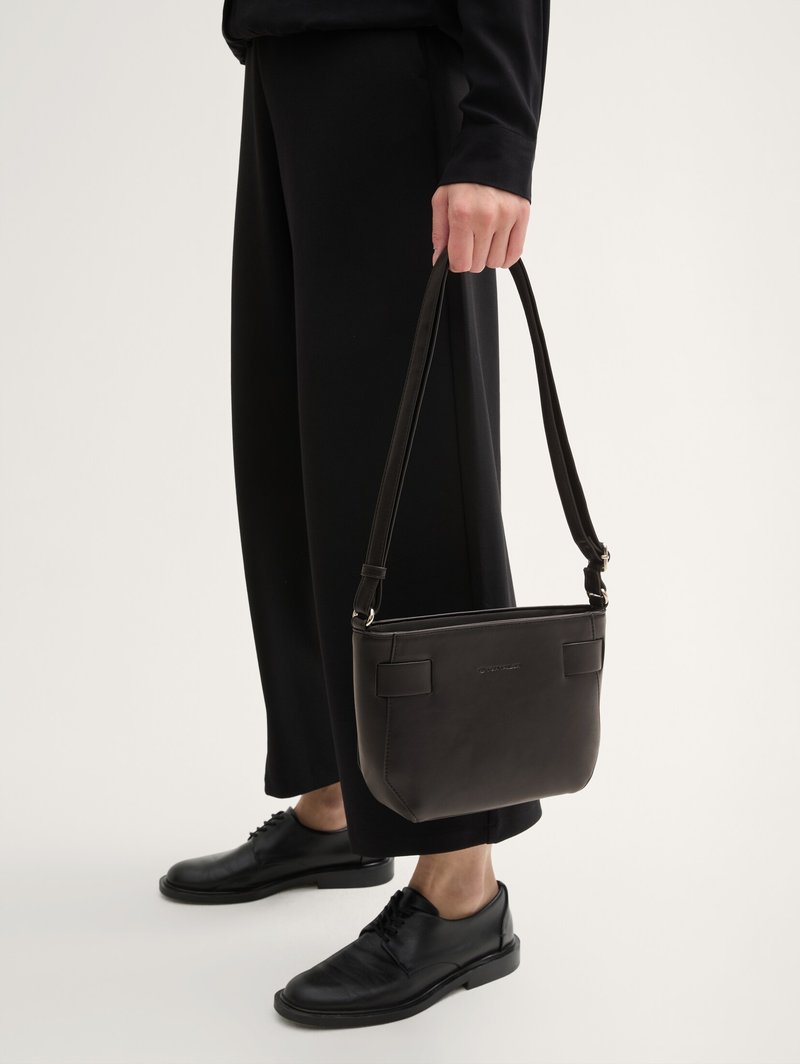 Personne vêtue de noir tenant un sac à bandoulière en cuir noir, portant un pantalon noir et des chaussures à lacets noires sur un fond neutre.