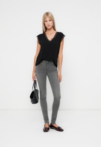 Blusa nera con pizzo sul colletto, abbinata a jeans aderenti grigi. La modella tiene in mano una borsa nera e indossa delle ballerine bordeaux.