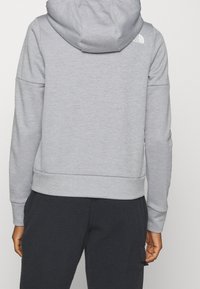 Persona que lleva una sudadera con capucha gris claro y pantalones de forro polar oscuros, vista desde atrás contra un fondo plano.