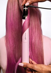 Ett pink hårstylingsverktyg lockar långt, rosa-tonat hår. Enheten har en elegant design med svarta accentdetaljer och texturerat grepp.