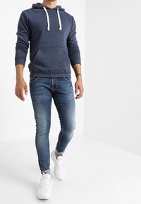 Felpa blu navy con lacci bianchi e tasca frontale, abbinata a jeans skinny azzurri e sneakers bianche. Modello in posa eretta.
