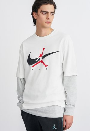 Valkoinen lyhyttä hihaista t-paitaa, jossa on punainen ja musta graafinen logo, jossa on hyppäävä hahmo, kerroksittain harmaan pitkähihasen paidan päällä. Mustat housut, joissa on logo.