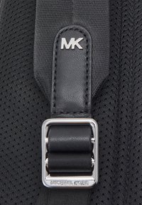 Tissu noir avec une surface texturée, comportant des accessoires en métal argenté. La sangle comprend un accent en cuir avec les initiales gravées "MK."