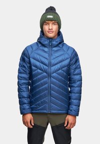 Niebieska kurtka puchowa w wzór zygzak, z wysokim kołnierzem i zapięciem na zamek. Noszona z oliwkowozieloną dzianinową czapką z pomponem.