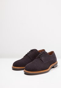 Chaussures à lacets en daim bleu foncé, avec une texture lisse, un bout arrondi, une semelle marron clair contrastante et des détails de couture minimalistes.