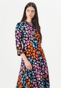 Cvečni plašč z črnim ozadjem, ki ima pisane vzorce v oranžni, roza in modri barvi, z gumbnim zapiranjem spredaj in rokavi do tri četrtine.