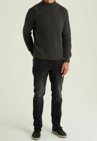 Grauer gestrickter Pullover mit hohem Kragen und strukturiertem Muster. Kombiniert mit dunklen Jeans und grünen Turnschuhen, stehend vor hellem Hintergrund.