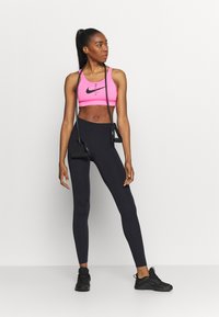 Sujetador deportivo rosa con el logo negro de Nike, combinado con leggings negros de talle alto. La modelo lleva zapatillas deportivas negras y sostiene un bolso pequeño.