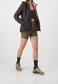 Veste noire imperméable avec doublure rose, soutien-gorge de sport marron, short vert et chaussures de trail multicolores avec semelles orange et chaussettes noires.