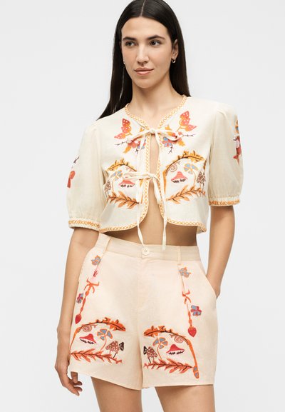Νεαρή γυναίκα φοράει εκρού κέντητο crop top με μοτίβα πεταλούδας και λουλουδιών, σε συνδυασμό με σορτς ψηλόμεσα με παρόμοια σχέδια.