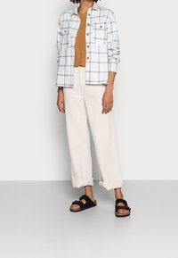 Witte geruite blouse met blauwe lijnen enbruine knopen, gecombineerd met een bruine top en crèmekleurige wijde broek, afgewerkt met zwarte sandalen.