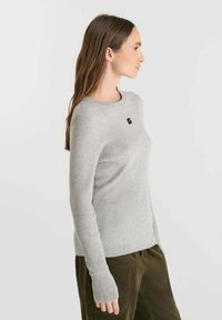 Light gray ribbed sweater with a crew neck and black logo detail on the chest. Long sleeves with extended cuffs. Soft texture. Casual fit.