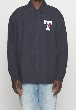 Mann trägt eine dunkelblaue Knopfjacke mit elastischen Bündchen und einem weißen „T“-Logo mit der Aufschrift „Tommy Jeans“ auf der Brust, kombiniert mit hellblauen Jeans.