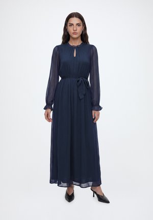 Femme portant une longue robe bleu marine avec des manches longues transparentes, un col en forme de trou de serrure, une ceinture à la taille et des talons noirs à bout pointu, debout sur un fond blanc.