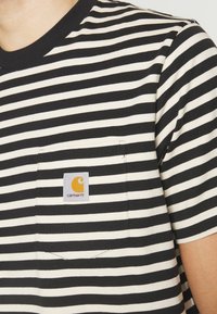 T-shirt rayé noir et crème avec une poche arborant un patch logo Carhartt. Le tissu semble lisse avec un design classique à col rond.