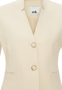 Béžový blazer se strukturálním designem, s klopovým límcem, dvěma kulatými zlatými knoflíky a bočními kapsami.