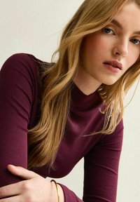 Burgundy ribbad polotröja med långa ärmar, kompletterad med ett gyllene armband på handleden och vågigt blont hår.