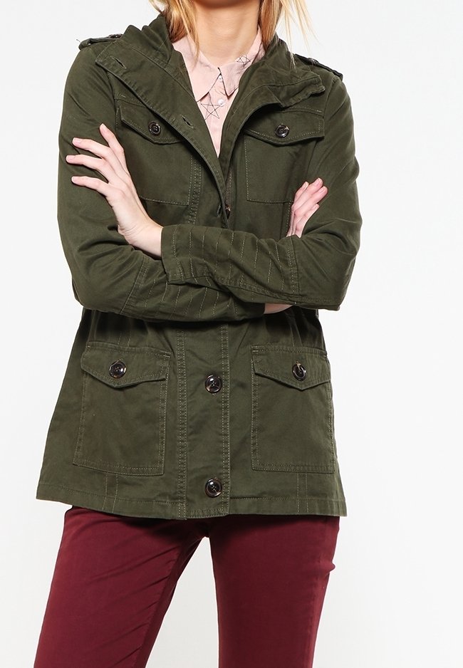 Osoba ubrana w oliwkową, zapinaną na guziki kurtkę z kołnierzem i kieszeniami klapowymi, w połączeniu z burgundowymi spodniami, stojąca z założonymi rękami.