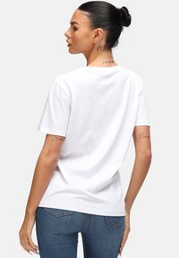 T-shirt en coton blanc à coupe décontractée, manches courtes et col ras du cou. Présente un logo discret au dos. Porté avec un jean bleu.