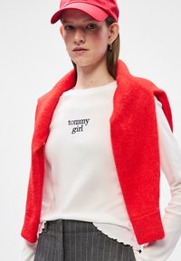 Pulover tricotat roșu așezat peste un top alb cu "tommy girl" în text negru; modelul poartă o șapcă roșie și pantaloni cu dungi.