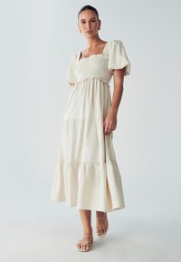 Bežinė vasarinė suknelė su kvadratiniais surišimais, pūstomis rankovėmis ir sluoksniuota sijonu. Išklota apatinė dalis ir derinama su nėriniuotais sandaliais.