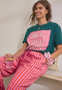 Leende kvinna sitter på en träbänk, klädd i en teal-färgad tröja med rosa text "BRING COFFEE," randiga rosa och orange byxor, håller ett glas med citronvatten.