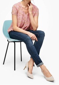 Blusa rosa chiaro con motivo floreale, jeans blu scuro e scarpe con tacco beige; seduta su una sedia blu chiaro.