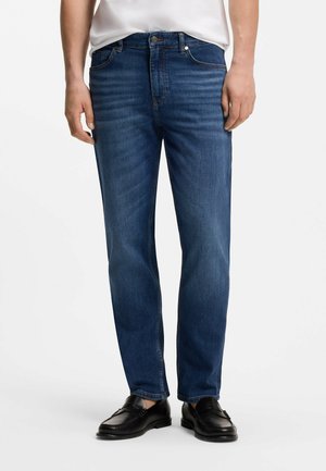 Mężczyzna ubrany w niebieskie proste jeansy, białą koszulę i czarne skórzane mokasyny, stoi na jednolitym białym tle.