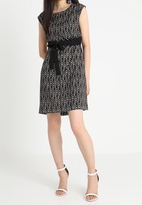 Černé šaty s kulatým vzorem, krátkými rukávy a páskem v oblasti pasu. Doplněné bílými sandály na podpatku. Hladká textilie.