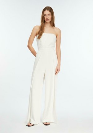 Kobieta o długich włosach, ubrana w białą, szeroką, bezramieniową kombinezon i czarne sandały na kwadratowym nosku, stojąca na jasnym, jednolitym tle.