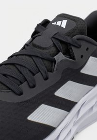 Svart sportskor med texturerat tyg, tonala skosnören, gummisula och tre distinkta silvriga ränder på sidan.